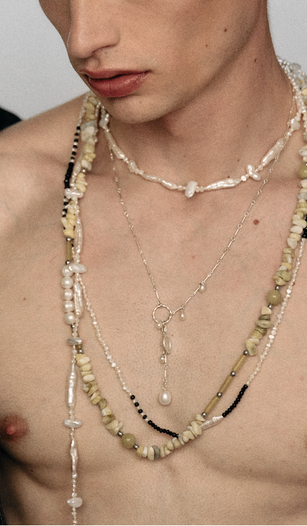 A close-up of a model's bare chest and lower face wearing multiple layered bohemian-style necklaces, including shell beads, pearls, and silver chains.